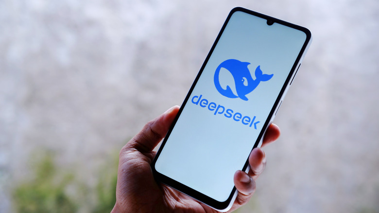 Рука с мобильным телефоном, отображающим имя DeepSeek и логотип. Фон изображения серый и размытый.