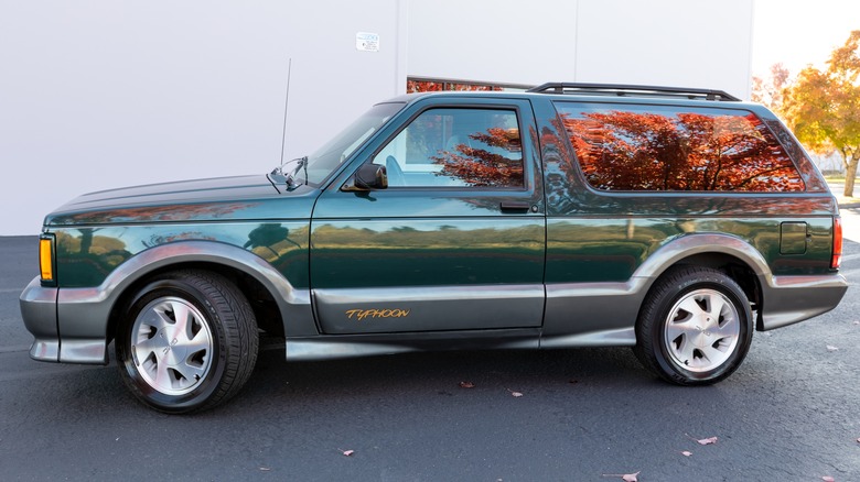 Боковой профиль блестящего зеленого на сером GMC Typhoon 1993 года, изображенного на белом здании, с оранжевыми листьями близлежащего дерева, отражающимися из окон.