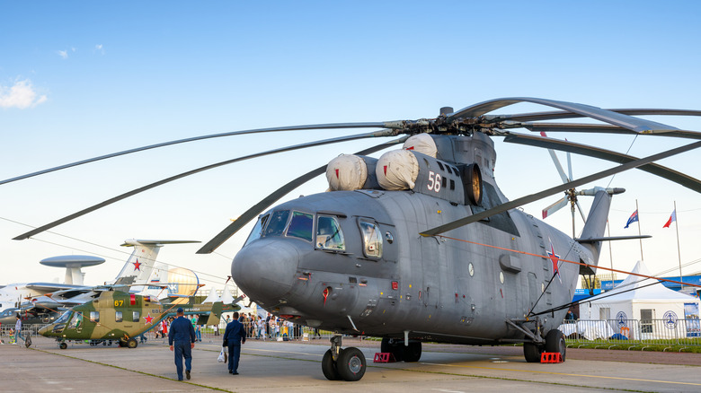 Mil Mi-26 Halo припарковался на стенде с двумя сотрудниками, осматривающими транспортное средство, видом на фронт-порт.