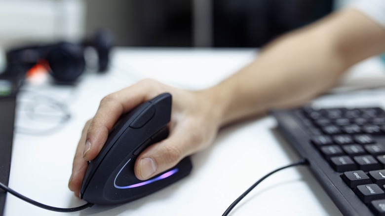 Рука, захватывающая черную вертикальную мышь на белом столе с аксессуарами для ПК на заднем плане