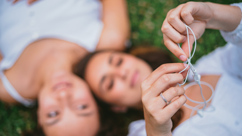 Два человека лежат в траве, один из которых держит белые проводные наушники с яблоками до камеры, чтобы распутать их.