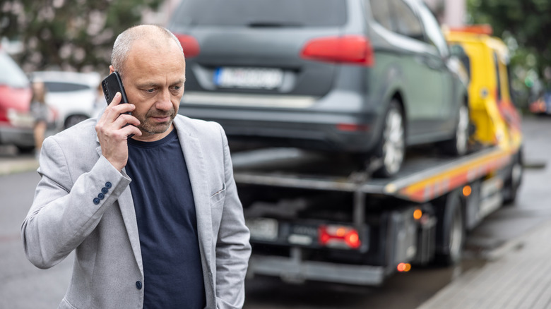 Человек среднего возраста, звонящий по телефону в качестве эвакуатора, несет машину сзади.