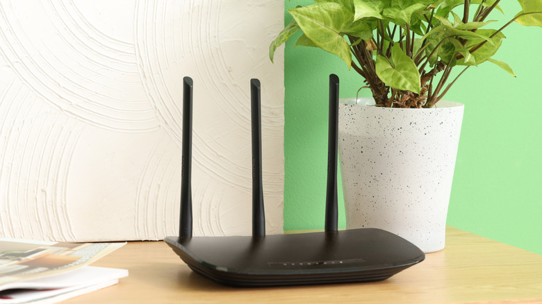 Черный маршрутизатор с тремя антеннами, торчащими на загроможденной поверхности растением.