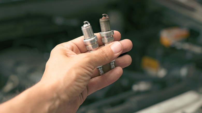 Механик, удерживающий новую и использованную свечу зажигания над отсекой двигателя.