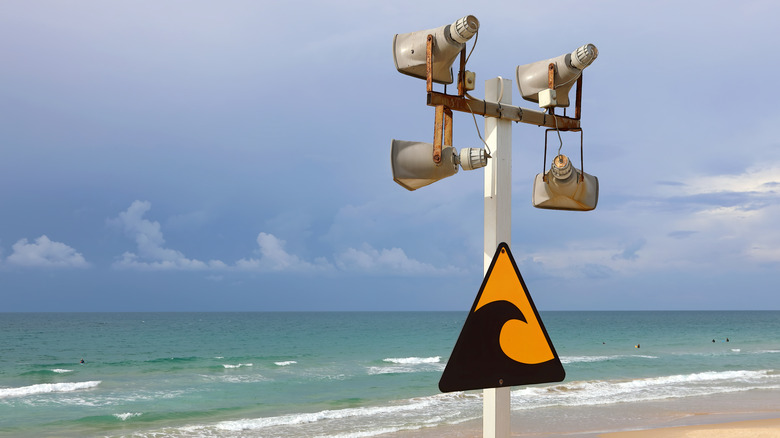 Знак, предупреждающий о цунами, на столбе с динамиками и пляжем на заднем плане.