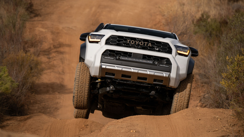 Белый внедорожник мчится через большие неровности по песчаной тропе, поросшей кустарником по обе стороны. Видна его ходовая часть