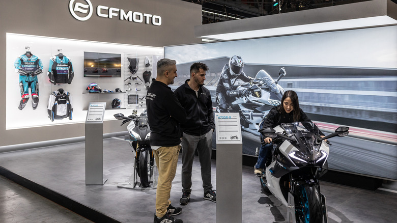 Стенд и подиум CFmoto, окрашенные в серый цвет, на закрытом торговом мероприятии, где люди рассматривают их предложения.