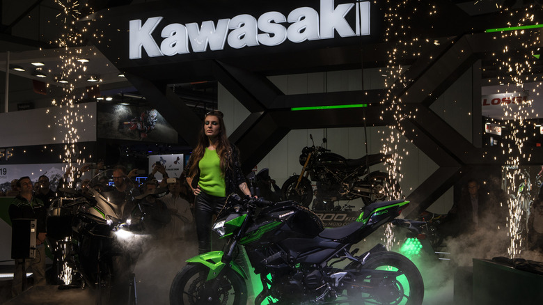 Мотоцикл Kawasaki Versys 1000 представлен на выставке в темноте под освещенным логотипом Kawasaki.