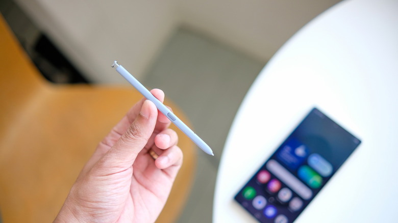 Рука, держащая стилус над телефоном, сидящим на белом столе