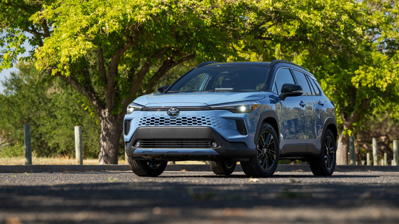 Toyota Corolla Cross Hybrid 2026 года припаркована на обсаженной деревьями улице.