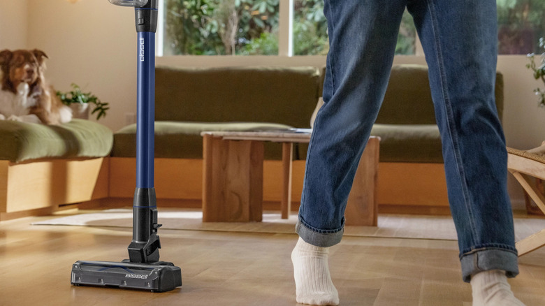 Человек в джинсах и носках идет рядом с беспроводным пылесосом Bissell, на заднем плане на диване сидит собака.