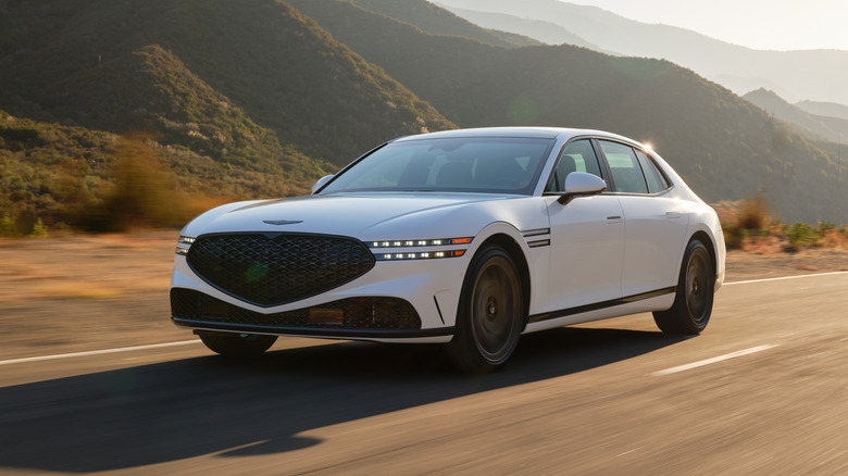 Белый Genesis G90 2026 года едет по дороге, вид спереди в 3/4, на заднем плане горы