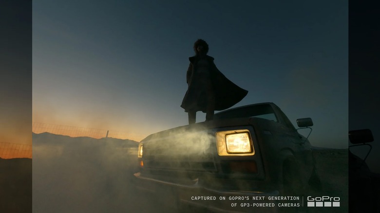 Человек, стоящий на капоте автомобиля с горящими фарами в сумерках.