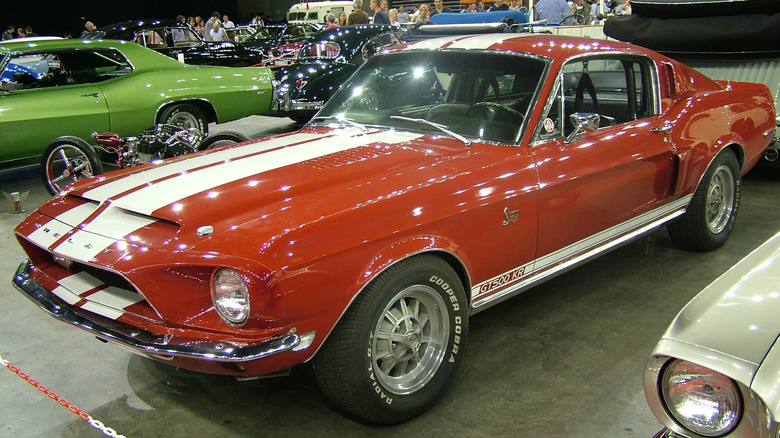 Ford Mustang Shelby GT500 KR 1968 года, припаркованный в помещении среди классических автомобилей и мотоциклов, вид спереди слева в 3/4