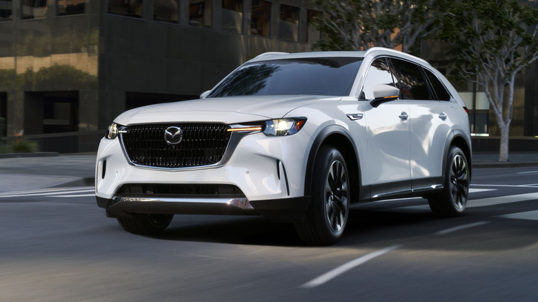 Mazda CX-90 PHEV на скорости в городе, вид спереди на 3/4, белый кузов