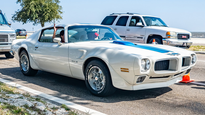 Вид спереди на 3/4 бело-синего Pontiac Trans Am начала 1970-х годов, едущего рядом с пляжем.