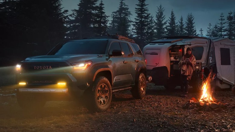 Зеленая Toyota 4Runner с включенными фарами, буксирующая кемпер, припаркованная рядом с костром.