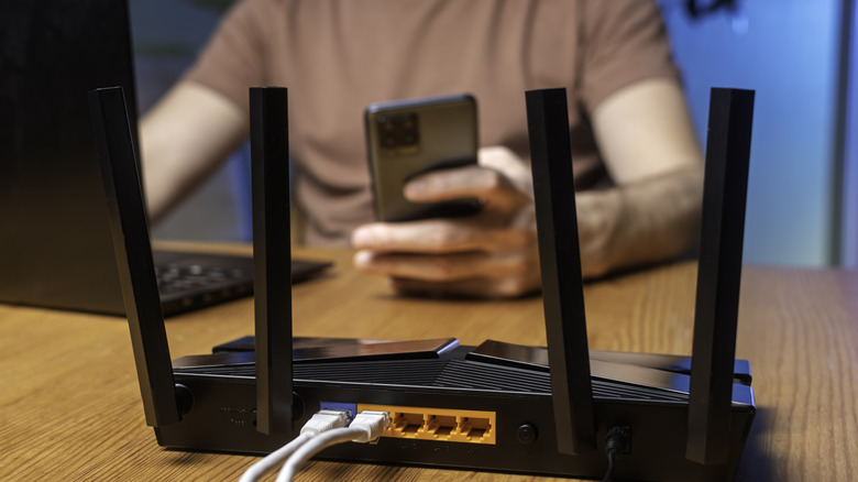 Черный Wi-Fi-маршрутизатор размещен на столе, и мужчина пользуется ноутбуком и смартфоном