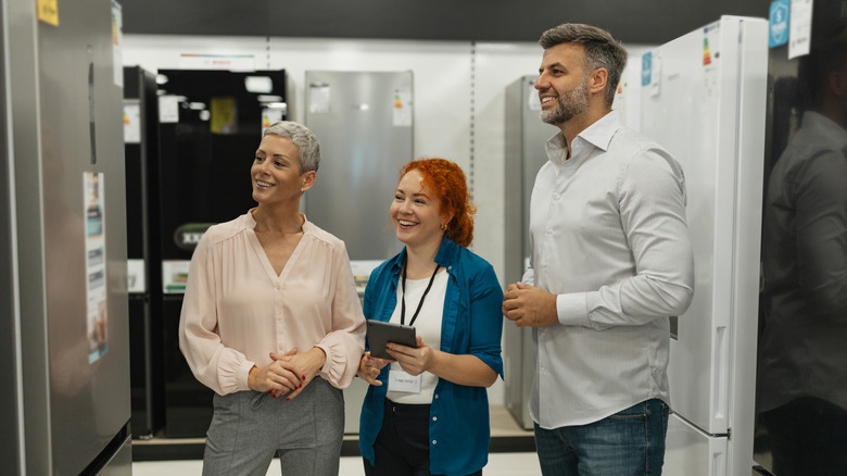Улыбающаяся пара смотрит на холодильник вместе с улыбающейся продавщицей.
