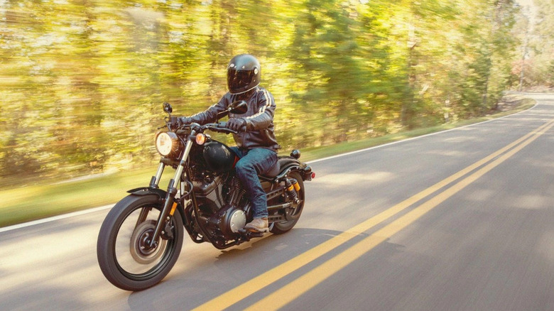 Гонщик в шлеме, закрывающем все лицо, едет на велосипеде Yamaha Bolt R-Spec 2026 года выпуска по лесной дороге, на которой видны двойные желтые линии.