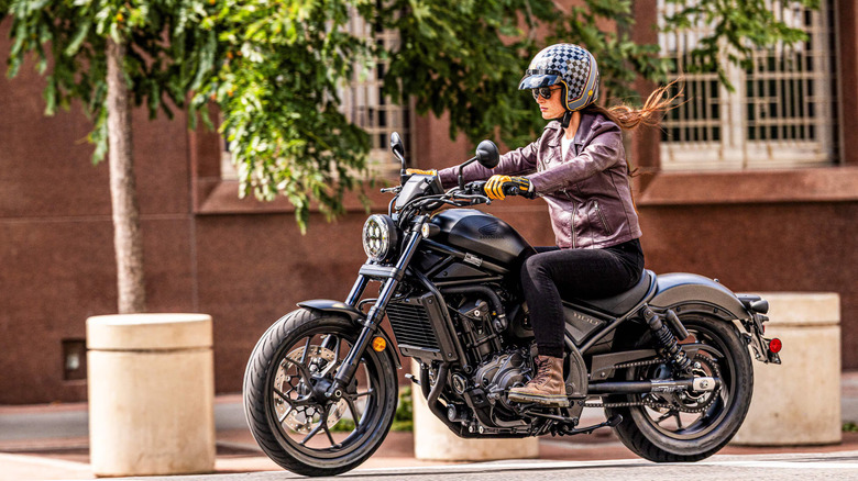 Женщина-гонщик в полной экипировке, шлеме и ботинках едет на Honda Rebel 1100 2026 года выпуска по пригородным улицам.