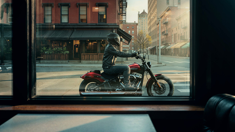 Мужчина-гонщик в кожаной экипировке и шлеме на красном Harley Street Bob, сфотографирован из проезжающей мимо витрины магазина.