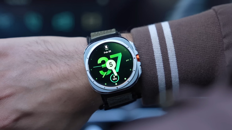 Рука в одежде с длинными рукавами и часами Galaxy Watch Ultra первого поколения с видимым неосовременным циферблатом.