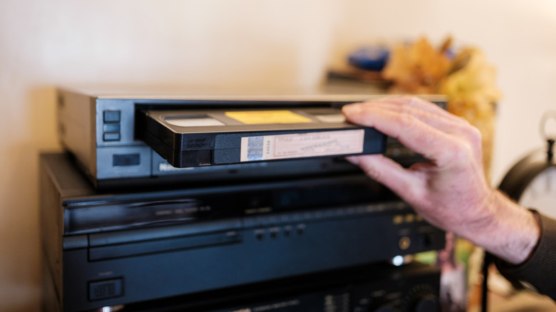 Рука вставляет кассету VHS в черный видеомагнитофон