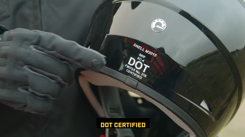 Палец в перчатке указывает на сертификационную этикетку DOT на задней части мотоциклетного шлема.