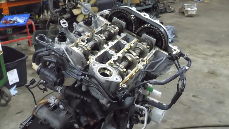 Разобранный 1,0-литровый двигатель Ford EcoBoost стоит на стенде в оживленной мастерской, а его клапанный механизм, распределительные валы и компоненты ГРМ полностью выставлены напоказ под освещением цеха.