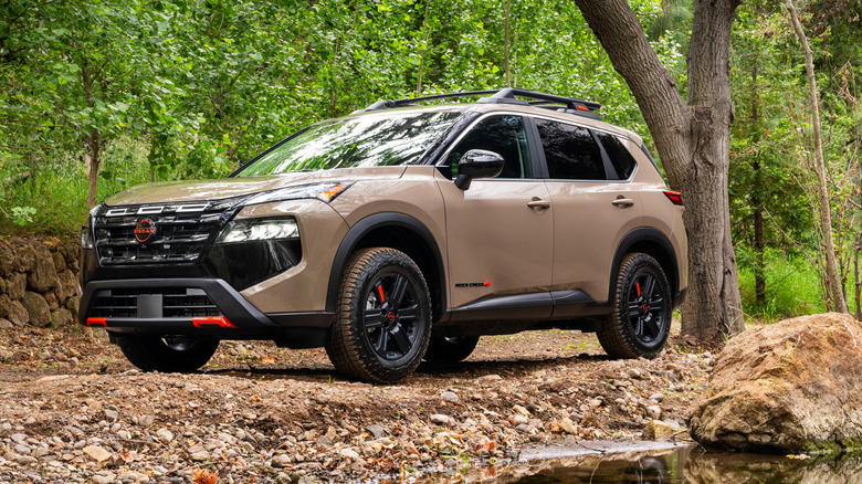 Вид спереди на 3/4 коричневых внедорожников Nissan Rogue 2026 года выпуска, припаркованных возле ручья.