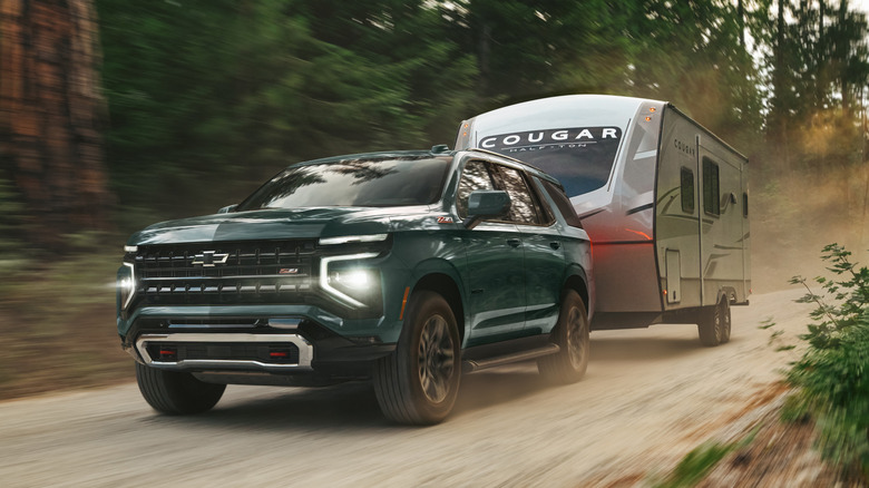 Вид спереди на 3/4 зеленого Chevy Tahoe 2026 года, буксирующего прицеп по грунтовой дороге.