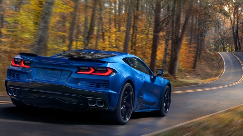 Вид сзади на синий Chevrolet Corvette E-Ray 2026 года, едущий по лесной двухполосной дороге
