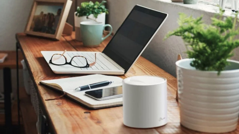 Маршрутизатор TP-Link Mesh System стоит на деревянном столе рядом с растением в горшке, открытым ноутбуком и другими офисными предметами.