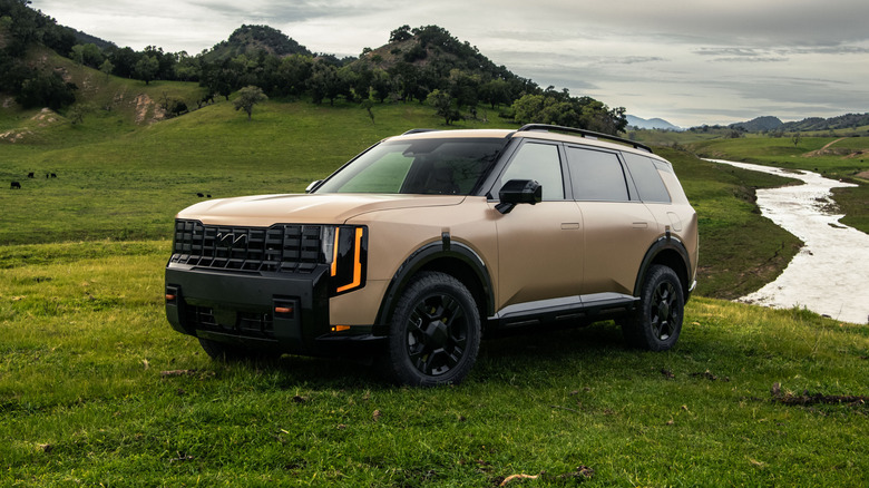 Коричневый Kia Telluride 2027 года выпуска, припаркованный на траве возле холмов и реки.