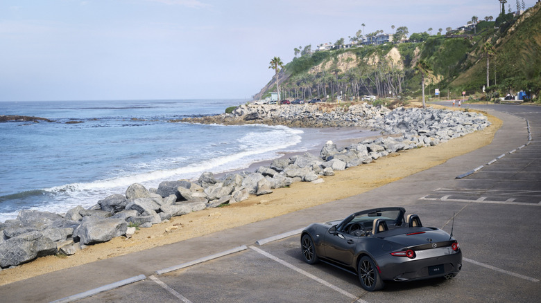 Mazda MX-5 2024 года припаркована перед каменистым пляжем, вид сзади слева в 3/4
