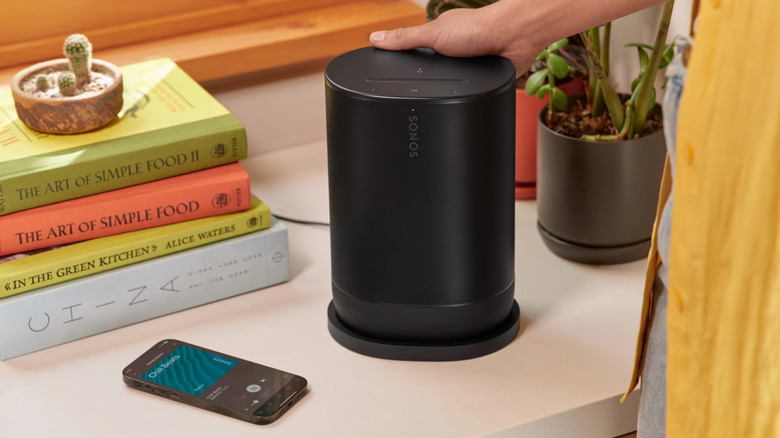 Человек, касающийся портативной колонки Sonos Move 2, окруженный книгами и смартфоном.