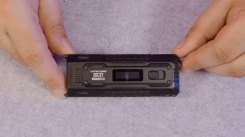 Руки сверху вниз держат фонарик Nitecore EDC37 на сером фоне.