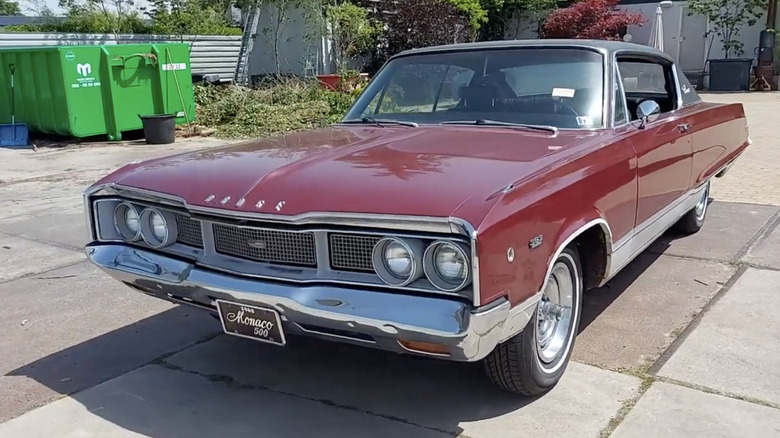 Купе Dodge Monaco 500 1969 года с жесткой крышей, окрашенное в темно-красный металлик, стоит на солнечной подъездной дорожке.