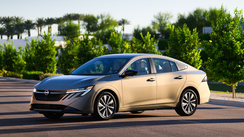 Золотой Nissan Sentra 2027 года выпуска припаркован на тротуаре на фоне рядов деревьев.