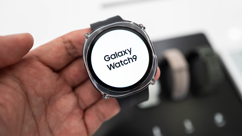 Рука держит Samsung Galaxy Watch 9, название которого отображается на белом экране.